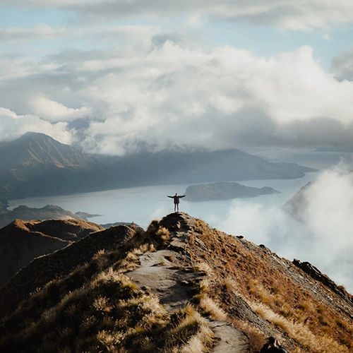 A distant view of a mountain peak and a man standing backwards with his arms raised, looking straight ahead.